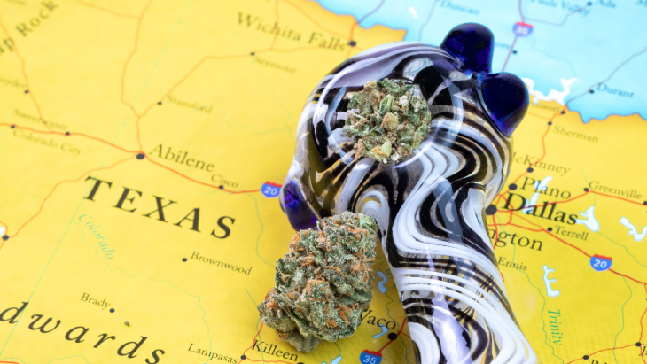 A glass pipe filled with cannabis and a cannabis bud rest atop a map of Texas featuring the city of Dallas