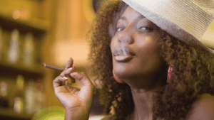 A Black woman with shoulder length curly hair wears a straw fedora, hoop earrings, and holds a legal cannabis blunt while exhaling smoke