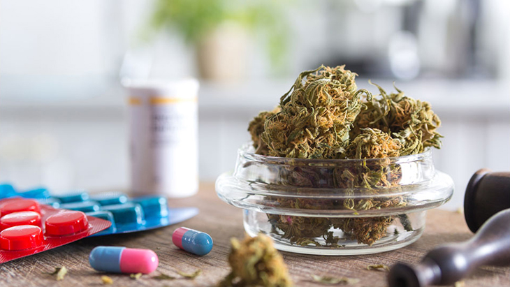 A small glass bowl of cannabis buds sits on a table next capsules of medicine