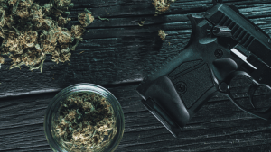 A small glass jar of legal cannabis flower sits on a table next to a black handgun
