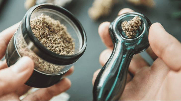 A closeup of hands holding a grinder and a glass pipe containing cannabis