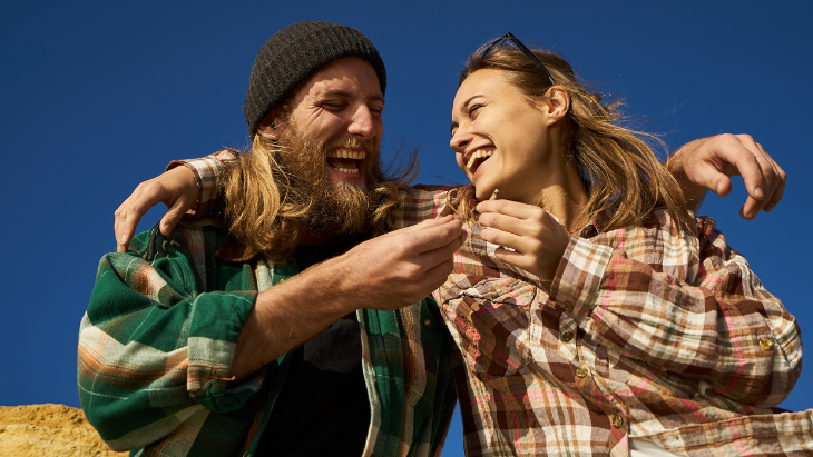 a man and woman stand outdoors with their arms around each other's shoulders smiling and laughing as she passes a cannabis joint to him
