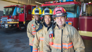three smiling firefighters stand in front of two firetrucks inside the firehouse