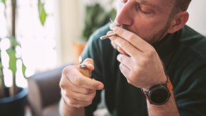 A man sits on his couch in a sunlit room and lights a legal cannabis joint