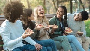 A group of smiling teens eats pizza while seated outdoors on a park bench